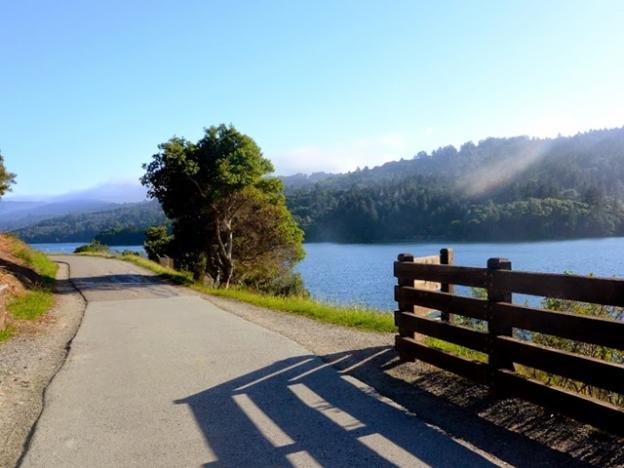 Paved path along Crystal Springs Trail with lake and wooded hills in the background on a sunny day.